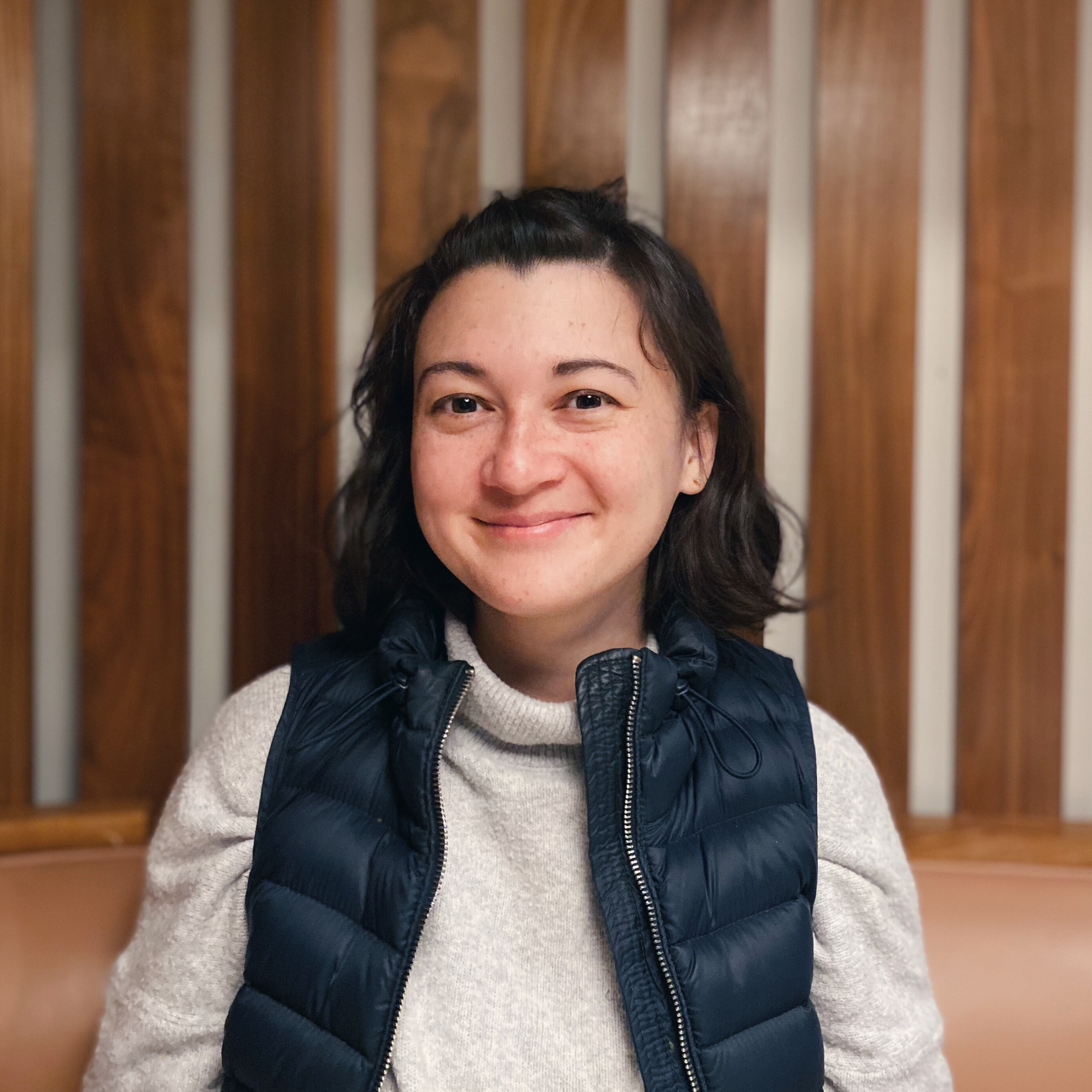 Becca sits in a booth with wood slats behind her. She is wearing a navy vest and grey sweater, and smiling.
